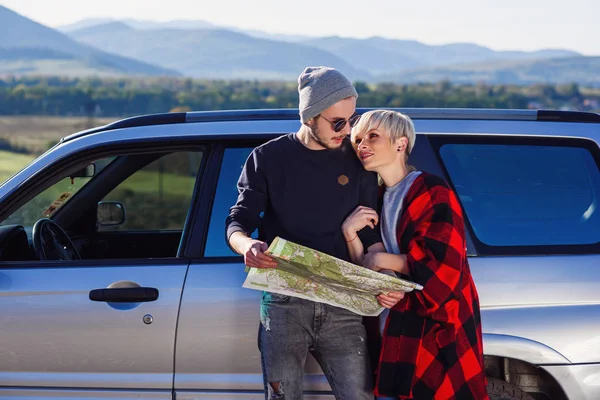 Casal turístico feliz com mapa de papel perto de carro alugado. Jovens na moda usando mapa. Viajar de carro em férias de verão em montanhas. Hipster homem e mulher tendo viagem ao ar livre . — Fotografia de Stock