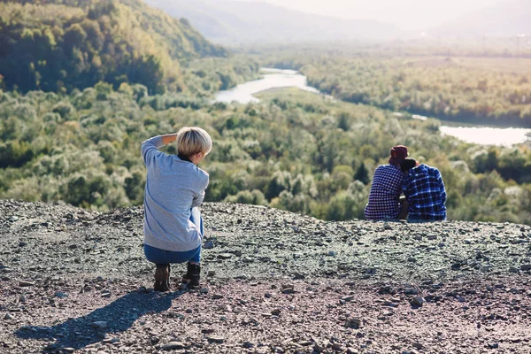 Jeune fille blonde photographe fait une photo de hipster homme et fille assis ensemble sur le sommet de la montagne au coucher du soleil. Voyages, tourisme et relations concept . — Photo