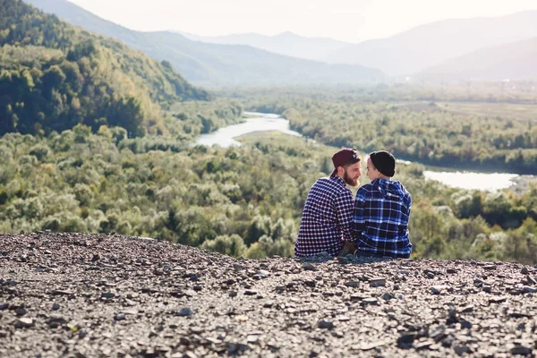 Ungt par i kärlek reser tillsammans i bergen. Glad hipster mannen och flickan sitter tillsammans på toppen av berget vid solnedgången bakgrund. Resor, turism och relationer koncept. — Stockfoto