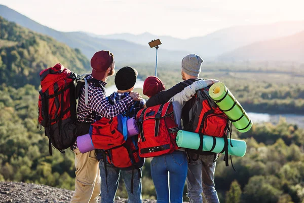 Group of young smiling people traveling together in mountains. Happy hipster travelers with backpacks making selfie. Traveling, tourism and friendship concept. View from back. Landscape background. — Stock Photo, Image