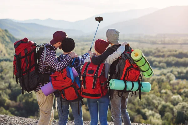 Group of young smiling people traveling together in mountains. Happy hipster travelers with backpacks making selfie. Traveling, tourism and friendship concept. View from back. Landscape background. — Stock Photo, Image