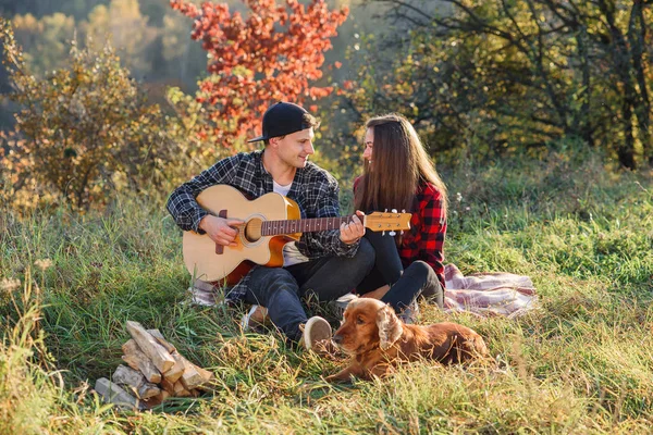 Lyckliga kaukasiska par med gitarr och deras hund har resten på picknick i parken på gräsmattan. — Stockfoto