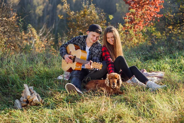 Happy lovely couple in casual clothes with guitar and their dog having rest on picnic in the park. — Stock Photo, Image