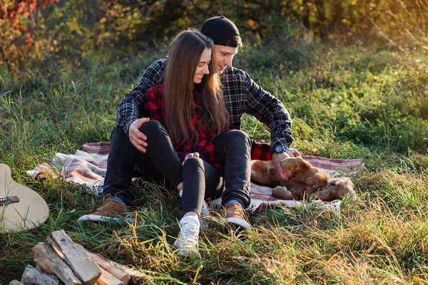 Feliz pareja caucásica con guitarra descansando en el picnic en el parque de primavera. Joven hombre y su novia jugando con su perro divertido . — Foto de Stock