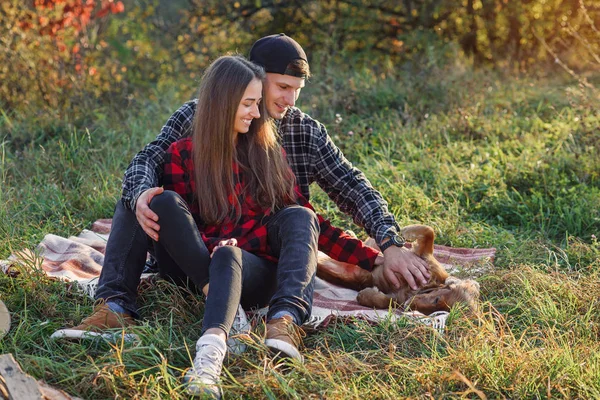 Feliz pareja caucásica con guitarra descansando en el picnic en el parque de primavera. Joven hombre y su novia jugando con su perro divertido . — Foto de Stock