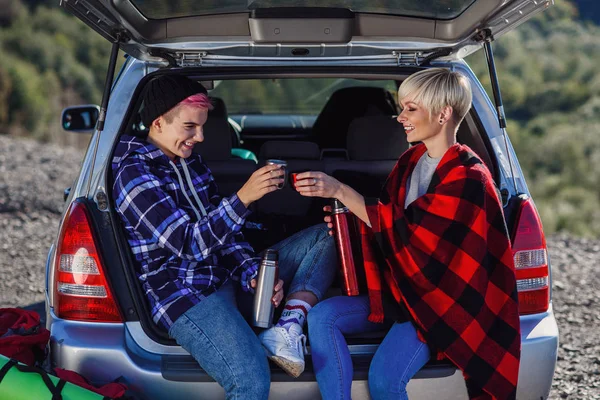 Viajar, turismo e conceito de amizade. Jovens amigos viajantes sentados no porta-malas do carro e bebem chá quente. Duas meninas hipster felizes desfrutando de férias nas montanhas . — Fotografia de Stock