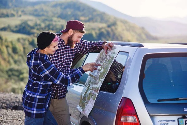 Casal turístico feliz com mapa de papel perto de carro. Jovens sorridentes usando mapa. Viajar de carro alugado em férias de verão. Hipster homem e mulher tendo viagem ao ar livre nas montanhas . — Fotografia de Stock