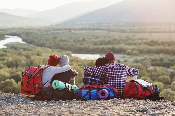 Group of young friends traveling together in mountains. Happy hipster travelers with backpacks sitting on the top of mountain at sunset background. Traveling, tourism and friendship concept. — Stock Photo, Image