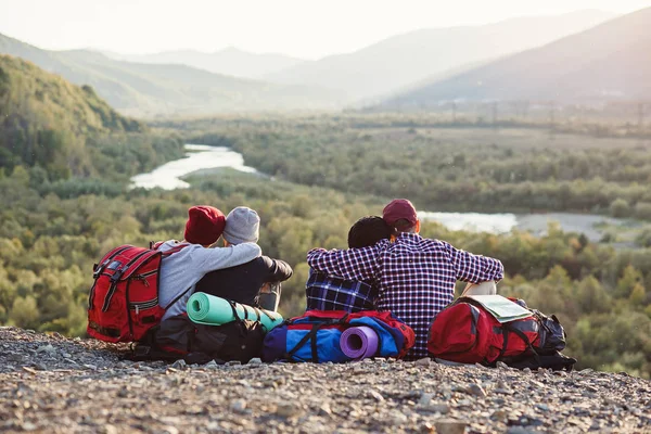 Group of young friends traveling together in mountains. Happy hipster travelers with backpacks sitting on the top of mountain at sunset background. Traveling, tourism and friendship concept. — Stock Photo, Image