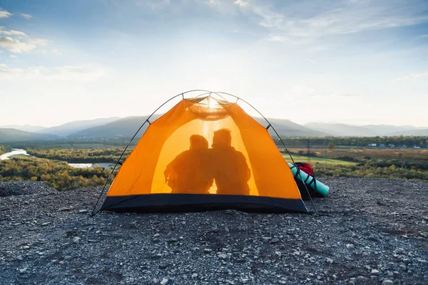 旅行、旅游和关系的概念。日落时分, 一个年轻人和他的女朋友在山顶上的橙色帐篷里互相亲吻的剪影. — 图库照片