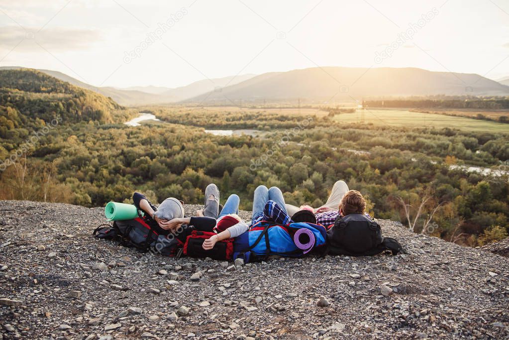 Traveling, tourism and friendship concept. Group of young friends traveling together in mountains. Happy hipster travelers with backpacks lying on the top of mountain at sunset background.