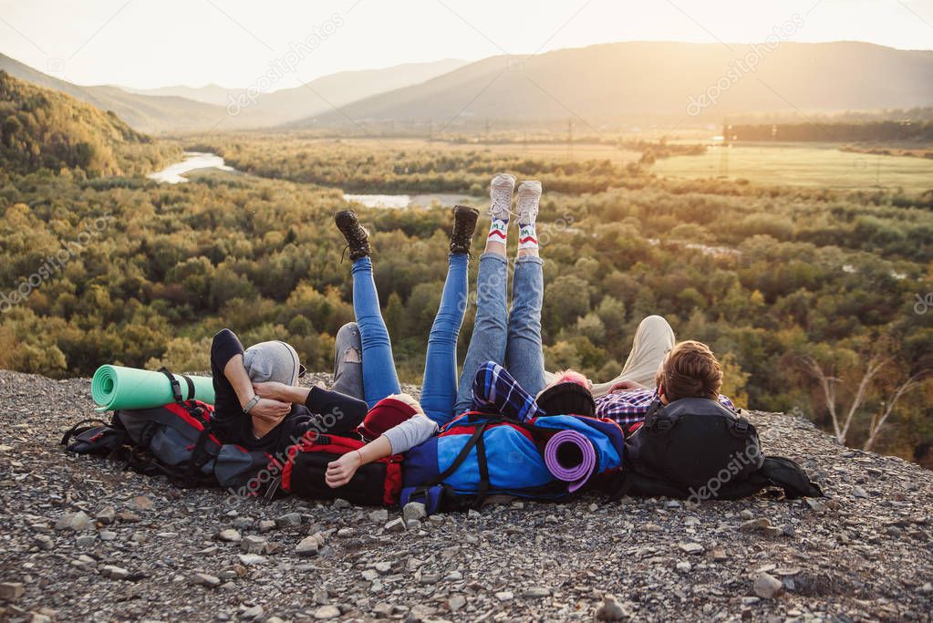 Traveling, tourism and friendship concept. Group of young friends traveling together in mountains. Happy hipster travelers with backpacks lying on the top of mountain at sunset background.