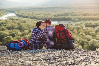 Birlikte dağlarında seyahat sırt çantaları ile genç çift. Mutlu hipster adam ve kız birlikte Dağı'nda günbatımı zemin üstünde oturuyor. Seyahat, turizm ve ilişkiler kavramı.