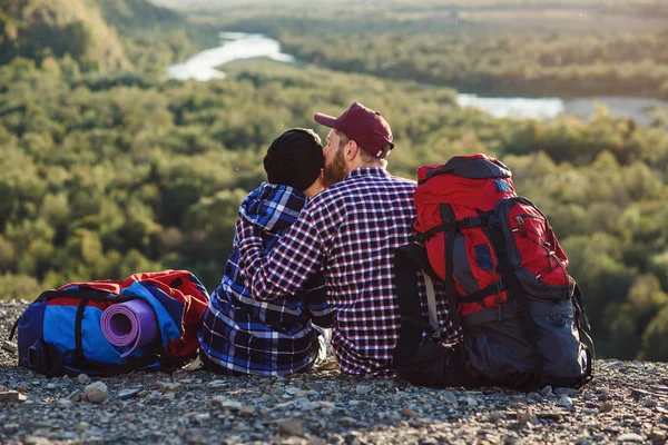 Молода пара з рюкзаками подорожує разом у горах. Щасливий хіпстер і дівчина сидять разом на вершині гори на фоні заходу сонця. Концепція подорожей, туризму та відносин . — стокове фото