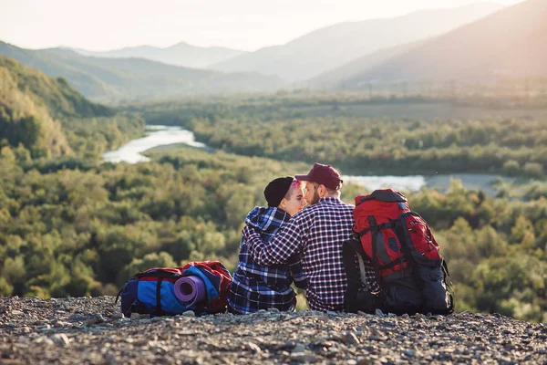 Giovane coppia con zaini che viaggiano insieme in montagna. Felice hipster uomo e ragazza seduti insieme sulla cima della montagna sullo sfondo del tramonto. Viaggiare, turismo e relazioni concetto . — Foto Stock