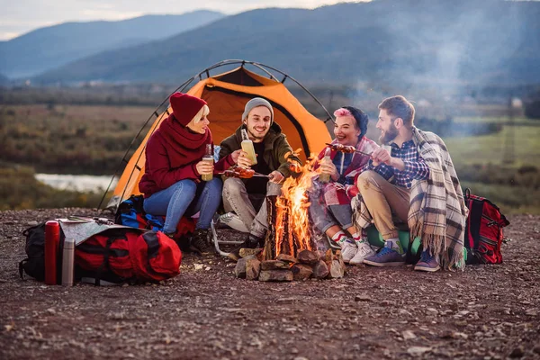 Companhia de jovens amigos fazendo um piquenique nas montanhas, eles estão conversando, rindo, bebendo bebida energética e assar salsichas na fogueira. Conceito de felicidade, juventude e gozo da vida . — Fotografia de Stock