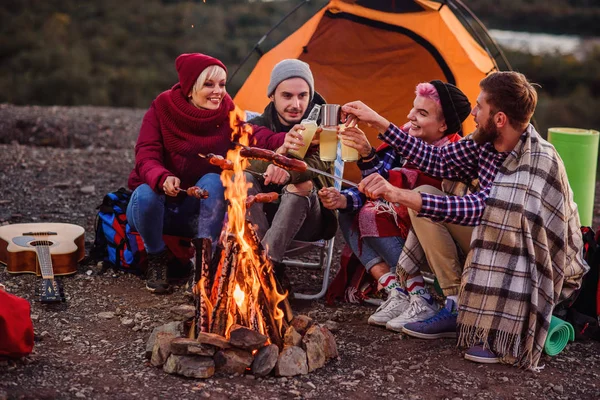 Company of young friends having a picnic by the mountains, they are chatting, laughing, drinking energy drink and bake sausages on bonfire. Concept of happiness, youth and enjoyment of life. — Stock Photo, Image