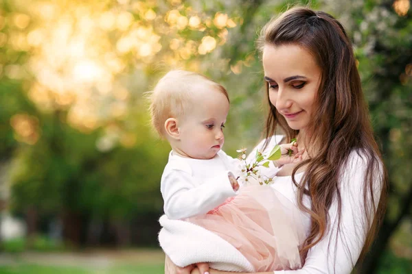 La mamma tiene la sua figlioletta tra le braccia tra gli alberi in fiore. Mamma e il suo piccolo bambino indossato rosa abito look familiare . — Foto Stock