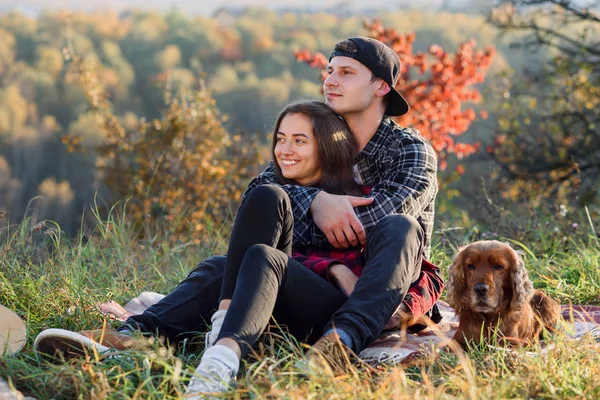 Felice coppia caucasica con il cane seduto sul prato nel parco primaverile. Giovane uomo e la sua ragazza godendo della natura . — Foto Stock