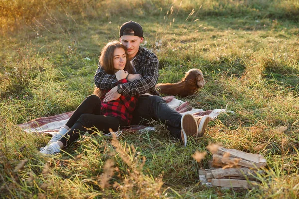 Young man hugs tenderly his pretty girlfriend. Young couple sits on the plaid at green lawn at sunset. — Stock Photo, Image