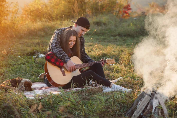 Ung man som spelar på gitarren för hans vackra flickvän. Lyckliga par med gitarr som vilar på picknick i vår park. — Stockfoto