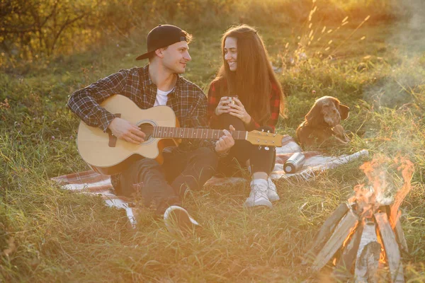 Young man playing on the guitar for his lovely girlfriend. Happy couple with guitar resting on picnic in spring park.
