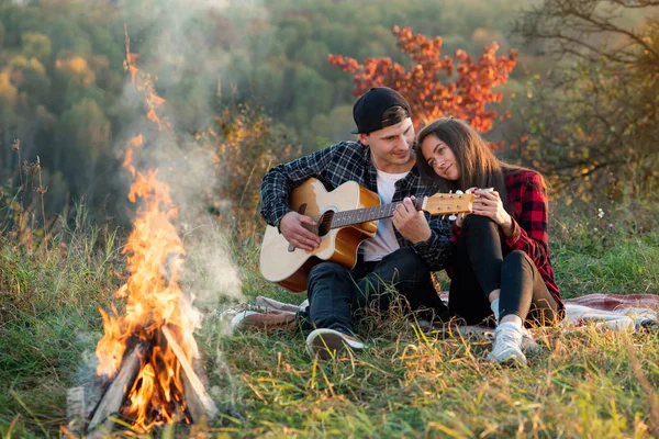 Young man playing on the guitar for his lovely girlfriend. Happy couple with guitar resting on picnic in spring park.