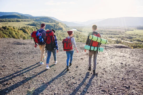 Vista desde atrás de cuatro amigos hipster con mochila de viaje cogidos de la mano y corriendo hacia la montaña en el fondo del atardecer . — Foto de Stock