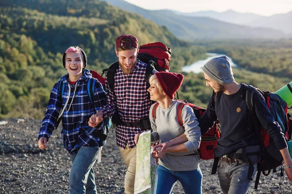 Group of four happy travelers are walking with the map and trying to find out the right way on mountain top at sunset, laughing and talking each other. — Stock Photo, Image