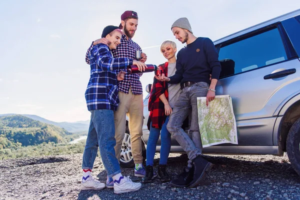 Summer travel concept. Happy friends using paper map near rented car in nature. Happy travelers in mountains on weekend vacation. Young woman pours hot tea into a cup for young man.