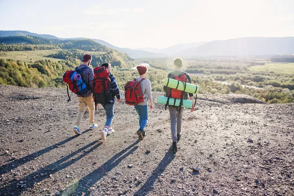 Vista desde atrás de cuatro amigos hipster con mochila de viaje cogidos de la mano y corriendo hacia la montaña en el fondo del atardecer . — Foto de Stock