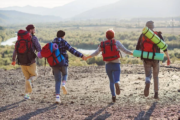 View from back of four hipster friends with travel backpack holding hands and running forward to the mountain on sunset background.