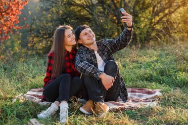 Genç mutlu hipster çift, bahar parkında çimlerde otururken akıllı telefon kullanarak selfie çekiyor. Modern çiftler, doğada oturan arkadaşlarıyla bir video görüşmesi yaparlar..