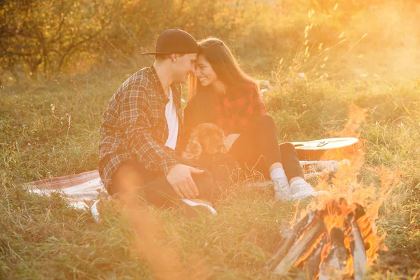 Happy caucasian couple in casual clothes with their funny dog sitting on the lawn in spring park. Young beautiful girl nd her handsome boyfriend leaned against each other at the nature. — Stock Photo, Image