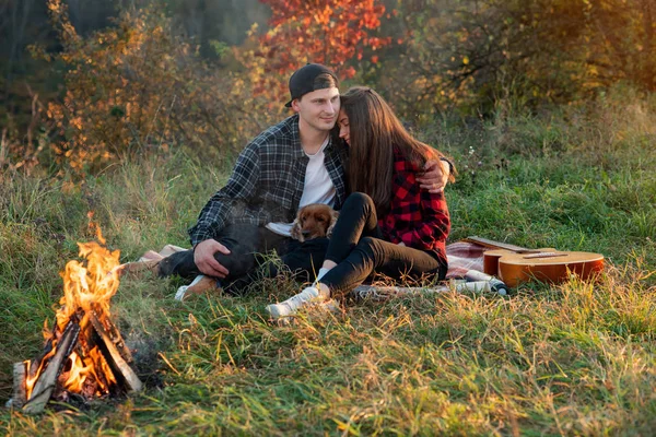 Feliz casal caucasiano com seu cão engraçado sentado no gramado no parque da primavera. Jovem menina bonita inclinou-se contra seu namorado e eles estão desfrutando na natureza . — Fotografia de Stock