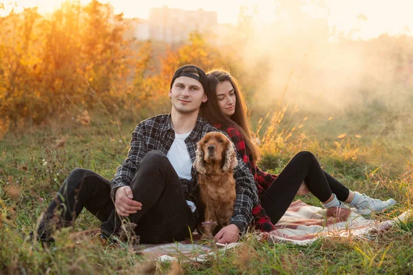 Casal caucasiano feliz em roupas casuais com seu cão descansando no gramado no parque da primavera. Jovem menina bonita inclinou-se contra seu namorado e eles estão desfrutando na natureza perto da fogueira . — Fotografia de Stock