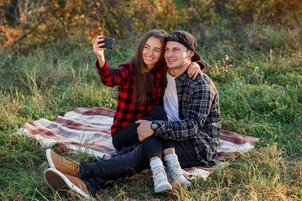 Jeune couple hipster heureux faisant selfie en utilisant un téléphone intelligent tout en étant assis sur la pelouse dans le parc de printemps. Élégant couple moderne ont un appel vidéo avec leurs amis assis dans la nature . — Photo