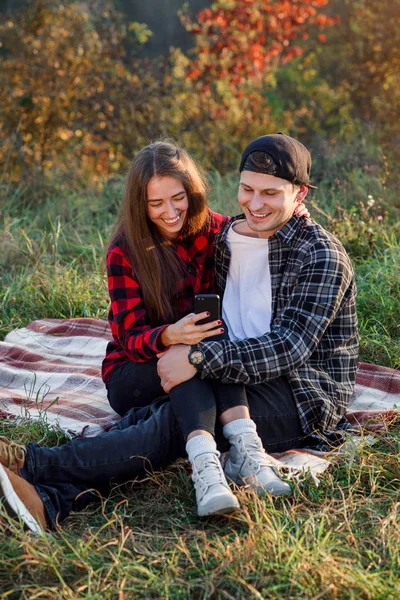 Bella ragazza si siede sul suo fidanzato in grembo e utilizzando smart phone sul prato nel parco. Hipster coppia moderna hanno una chiacchierata con i loro amici seduti nella natura . — Foto Stock