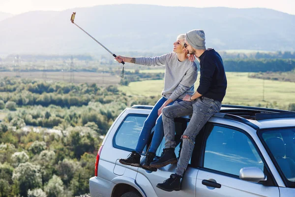 Jovem hipster casal no amor fazendo selfie usando telefone inteligente enquanto sentado no telhado de off-road carro no fundo da montanha . — Fotografia de Stock
