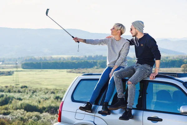Jovem hipster casal no amor fazendo selfie usando telefone inteligente enquanto sentado no telhado de off-road carro no fundo da montanha . — Fotografia de Stock