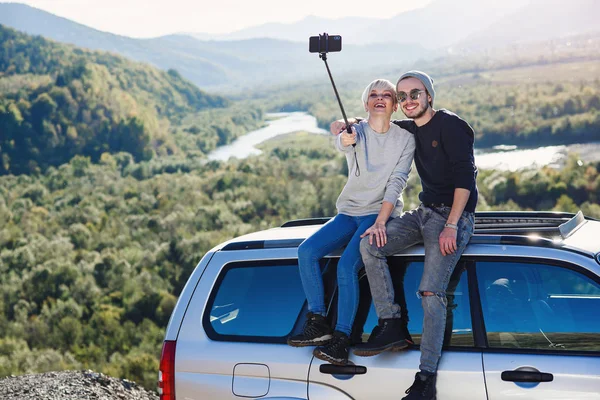 Jovem hipster casal no amor fazendo selfie usando telefone inteligente enquanto sentado no telhado de off-road carro no fundo da montanha . — Fotografia de Stock