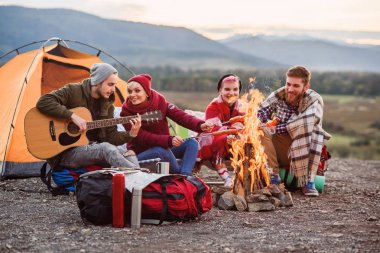 Mutlu arkadaşlar dağlarda piknik yapıyor, sohbet ediyorlar, enerji içecekleri içiyorlar, gitar çalıyorlar ve kamp ateşinde sosis pişiriliyorlar. Mutluluk kavramı, gençlik ve yaşam keyfi.