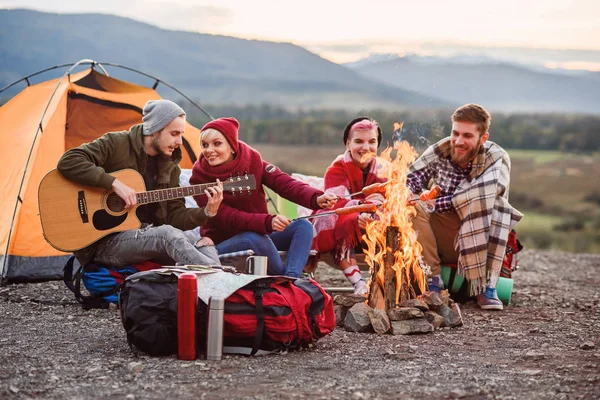 Amigos felices haciendo un picnic en las montañas, están charlando, bebiendo bebidas energéticas, tocando la guitarra y horneando salchichas en la fogata. Concepto de felicidad, juventud y disfrute de la vida . — Foto de Stock