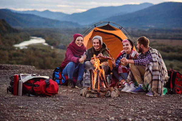 Company of young friends having a picnic by the mountains, they are chatting, laughing, drinking energy drink and bake sausages on bonfire. Concept of happiness, youth and enjoyment of life.