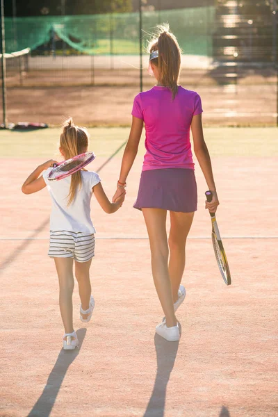 Kvinnlig tennis tränare håller sin lilla elev till händerna och de går samman efter träning på utomhustennisbana vid solnedgången. — Stockfoto