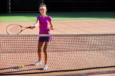 Açık tenis kortunda antrenman sırasında tenis topu servis eden şapkalı ve tenis formalı genç bir kadın. Spor giyimli güzel kız gün batımında tenis antrenmanı var..