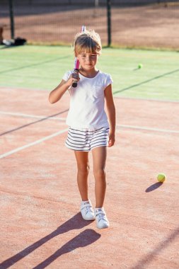 Küçük kız gün batımında açık tenis kortu üzerinde yürüyüş omzuna tenis raket ile beyaz üniforma giymiş.