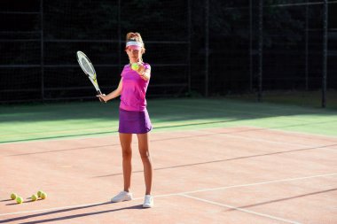 Açık tenis kortunda antrenman sırasında tenis topu servis eden şapkalı ve tenis formalı genç bir kadın. Spor giyimli güzel kız gün batımında tenis antrenmanı var..