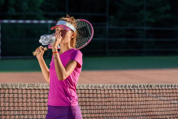 特写可爱的年轻女孩在运动帽饮用水从塑料瓶后硬网球训练在户外球场背景在日落. — 图库照片