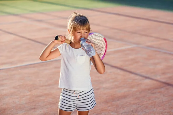 Gros plan drôle petite fille boire de l'eau de la bouteille après un entraînement de tennis dur au court extérieur au coucher du soleil. Sport, concept de santé . — Photo